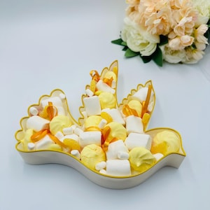 Può includere: Un piatto da portata a forma di colomba pieno di dolcetti gialli e bianchi, tra cui marshmallow e meringhe, accentuati da nastri arancioni. Il piatto ha una base color crema con un bordo dorato. Un bouquet di fiori color pesca e bianchi è sullo sfondo.