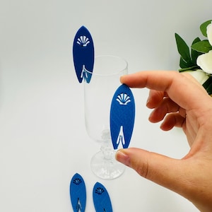 Könnte beinhalten: Blaue Surfbrett-Getränkeanhänger mit weißem Muscheldesign. Die Anhänger sind aus einem strukturierten Material gefertigt und zum Aufhängen am Glasrand bestimmt. Mehrere Anhänger sind zu sehen, einer wird von einer Hand gehalten.