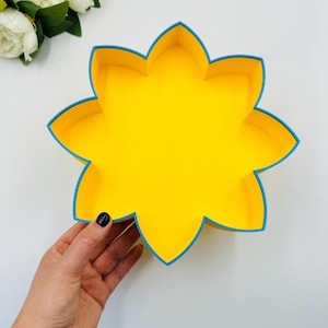 Puede incluir: Una bandeja amarilla en forma de flor con un borde azul. La bandeja está sostenida por una mano sobre un fondo blanco. Se ven flores blancas en la esquina superior izquierda.