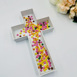 Op de afbeelding: Een wit, kruisvormig dienblad gevuld met kleurrijke snoepjes. De snoepjes zijn in verschillende kleuren, waaronder roze, geel, oranje, groen en bruin. Het dienblad staat op een witte ondergrond, met een boeket bloemen op de achtergrond.