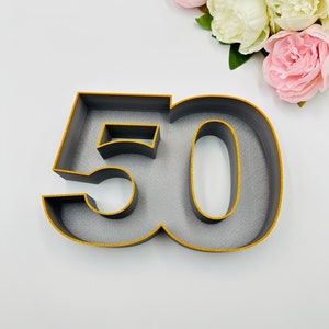 Könnte beinhalten: Ein graues Tortenaufsatz mit der Zahl 50 und goldenen Rändern. Die Zahl ist in einer fetten, blockartigen Schriftart gehalten. Der Hintergrund ist weiß mit rosa und weißen Blumen.