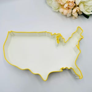 May include: A white serving dish shaped like the United States, with a bright yellow outline. The dish is empty and rests on a white surface. A bouquet of flowers is visible in the background.