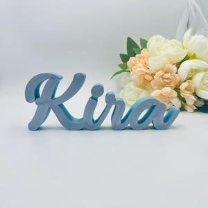 Könnte beinhalten: Ein hellblaues Holzschild mit dem Namen "Kira" in Schreibschrift. Das Schild steht vor einem Strauß weißer und pfirsichfarbener Blumen. Der Hintergrund ist weiß.