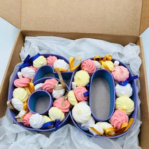 May include: A purple number 60 filled with colourful meringues in a cardboard box. The meringues are white, pink, and yellow, with blue and gold ribbons. The box is lined with white tissue paper.
