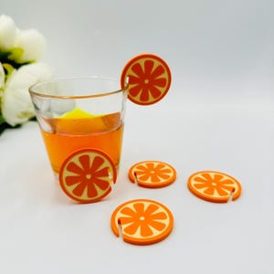 Puede incluir: Un vaso transparente lleno de líquido naranja, acompañado de posavasos con forma de rodajas de naranja. Los posavasos y la rodaja del vaso tienen un diseño naranja brillante con un centro naranja claro, creando una estética vibrante.