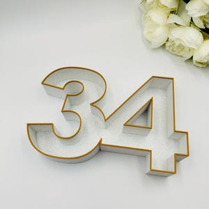 Könnte beinhalten: Eine dekorative Zahl 34 mit weiß gesprenkeltem Innenraum und goldfarbener Umrandung. Die Zahlen bestehen aus einem dicken Material, möglicherweise Holz oder MDF. Der Hintergrund ist weiß, mit einem Strauß weißer Blumen in der oberen rechten Ecke.