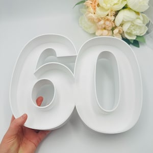 May include: A large white number 60, possibly for a birthday or anniversary. The number is hollow, designed to be filled. A bouquet of white and peach flowers is in the background.