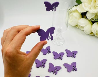 May include: Purple butterfly-shaped place card holders. The holders are made of a solid material and have a slot to hold a place card. A hand is holding one of the butterfly place card holders. A clear glass and white flowers are in the background.