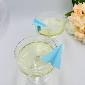 Pode incluir: Dois copos de cocktail transparentes cheios de um líquido amarelo claro. Cada copo é adornado com um avião de papel azul claro. Os copos têm um design canelado e estão sobre uma superfície branca. Um buquê de flores está no fundo.