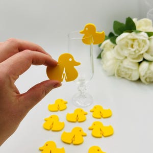 May include: Yellow rubber duck shaped drink markers. There are nine ducks in total, with one on a champagne flute and the rest on a white surface.