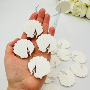 May include: A collection of white plastic seashell-shaped drink coasters. The coasters are arranged on a hand and a table.