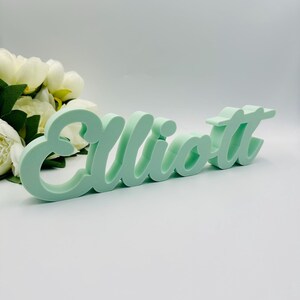 May include: A mint green wooden sign spells out the name "Elliott" in a cursive font. The sign is placed on a white surface, with a bouquet of white flowers in the background. The letters are thick and stand out against the background.