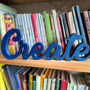 Könnte beinhalten: Ein marineblaues Holzschild mit der Aufschrift "Create" in Schreibschrift steht auf einem Holzregal vor einer Büchersammlung. Die Bücher haben verschiedene Farben und Größen und bilden einen lebendigen Hintergrund für das Schild.