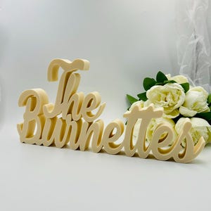 Könnte beinhalten: Ein cremefarbenes Holzschild mit der Aufschrift "The Burnettes" in einer Schreibschrift. Das Schild befindet sich vor einem Strauß weißer Blumen. Es ist ein dekorativer Artikel.