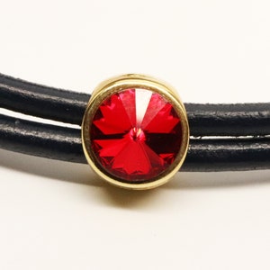 May include: A close-up of a red crystal bead with a gold setting. The bead is on a black leather cord.