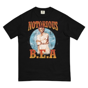 May include: Black t-shirt with a vintage-style graphic of a woman with blonde hair in front of a blue and white moon. The text "NOTORIOUS B.E.A." is printed in orange and yellow.