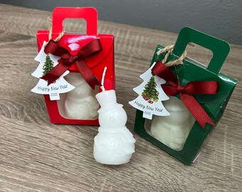 Könnte beinhalten: Zwei kleine Geschenkboxen mit durchsichtigen Fenstern, jede mit einer weißen Schneemann-Kerze. Die Boxen sind rot und grün mit einem roten Band und einem Etikett mit der Aufschrift "Frohes Neues Jahr 2023".