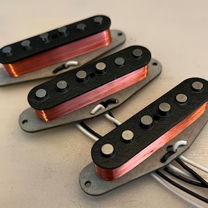 May include: Three guitar pickups with black tops, silver pole pieces, and copper wire coils. The pickups are mounted on grey metal plates with attached wires. The image is taken from a close-up angle.