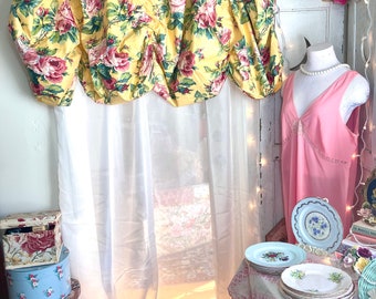 May include: A yellow floral valance with pink roses hangs over a white sheer curtain. The valance is gathered in the center and has a ruffled edge. The curtain is sheer and has a pink ruffle at the bottom. The window is behind the curtain and is letting in sunlight.