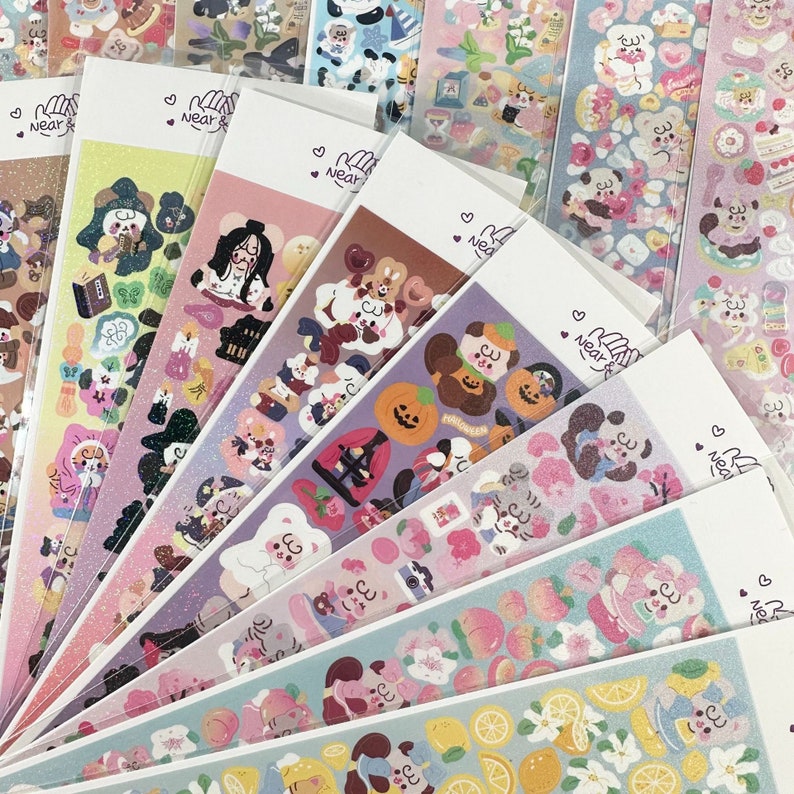 May include: A collection of colorful sticker sheets featuring various designs, including cute animals, food items, and seasonal themes. Each sheet has a white header with the text "Near &" and a heart symbol. The stickers are in clear plastic packaging.