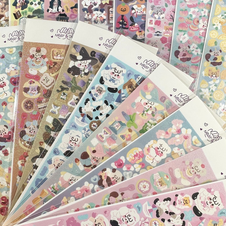 May include: A collection of sticker sheets with various designs, including cute characters, food items, and floral patterns. The stickers are in a variety of colors, including pink, blue, and gold. Each sheet has the text "Near & Dear" and a heart symbol.
