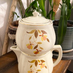 May include: Cream-colored ceramic teapot and sugar bowl set. The set features a floral design with brown, orange, and yellow accents, and gold trim. The teapot has a handle and spout, and the sugar bowl has a lid.
