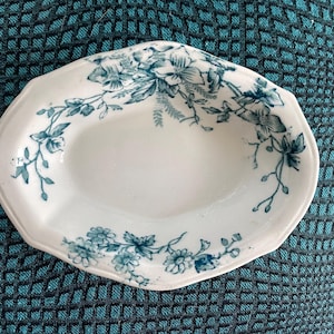 May include: White oval-shaped ceramic dish with a scalloped edge. The dish is decorated with a teal floral pattern around the rim. The floral design includes flowers and leaves. The dish has a smooth, glossy finish.