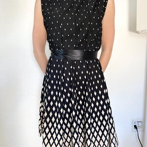 May include: A sleeveless black dress featuring a white diamond pattern that transitions into a larger grid design toward the hem. The dress includes a solid black satin-finish waistband and vertical pleats along the bodice and skirt.