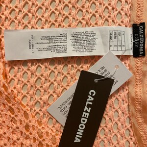 May include: A peach-colored garment with a textured, open-knit mesh pattern. Attached are white and black product tags featuring the brand name Calzedonia, size S, and manufacturing information including the text Made in Croatia and HR-13MA-1.