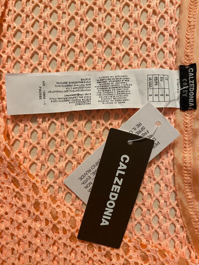 May include: A peach-colored garment with a textured, open-knit mesh pattern. Attached are white and black product tags featuring the brand name Calzedonia, size S, and manufacturing information including the text Made in Croatia and HR-13MA-1.