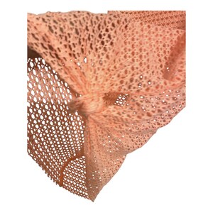 May include: A peach-colored fabric accessory featuring an open-knit, mesh-like texture. The material is gathered and secured with a central knot, creating a series of folds and drapes that highlight the intricate, repeating geometric pattern of the weave.