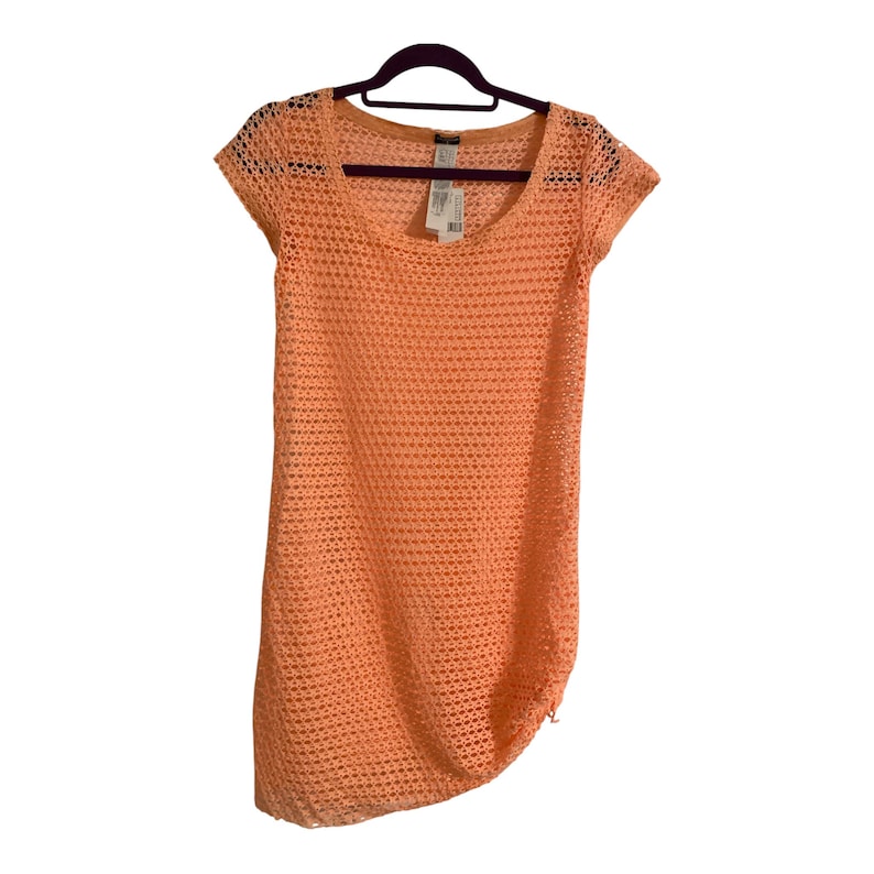 May include: A peach-colored, short-sleeved dress featuring an open-knit, mesh-like texture throughout. The garment has a wide, rounded neckline and is displayed on a dark-colored hanger, with a white clothing tag visible near the collar area.
