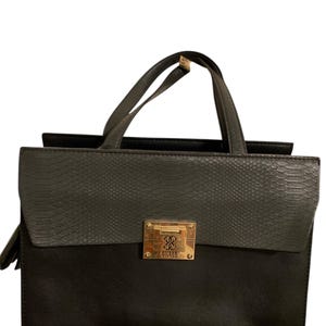 Könnte beinhalten: Eine schwarze und graue Handtasche mit rechteckiger Form. Die obere Klappe ist grau mit Schlangenhautstruktur, der untere Teil ist schwarz. Sie hat einen goldfarbenen Verschluss mit dem Markennamen "GUESS" und zwei schwarze Griffe.