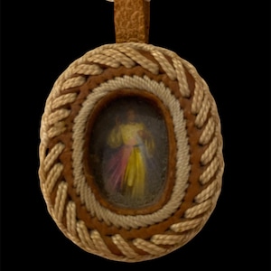 May include: Oval pendant with a religious image, featuring a braided rope design around the edges. The pendant has a leather backing and a cream-colored rope necklace. The image inside the pendant has yellow, pink, and purple hues.