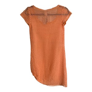 May include: A short-sleeved, peach-colored top featuring an open-knit, mesh-like crochet pattern throughout. The garment has a rounded neckline and a slightly asymmetrical hemline, constructed from a lightweight, semi-sheer textile material.