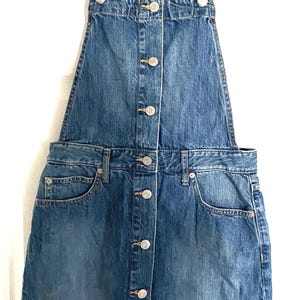 Op de afbeelding: Een blauwe spijkerbroekjurk met verstelbare bandjes en een knoopsluiting aan de voorkant. De jurk heeft twee voorzakken en hangt aan een zwarte hanger. Het kledingstuk is gemaakt van een medium-wash denim stof.