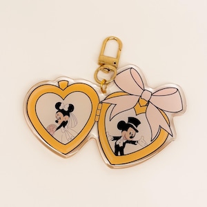 May include: A heart-shaped keychain featuring Mickey and Minnie Mouse in wedding attire. The keychain has a gold-colored clasp and a pink bow. The images of Mickey and Minnie are inside heart-shaped frames.