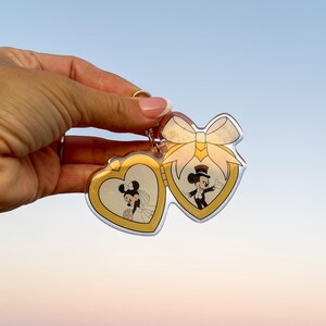 May include: A gold-colored heart-shaped keychain featuring Mickey and Minnie Mouse in wedding attire. The keychain has a bow design and a gold border. The background is a clear blue sky.