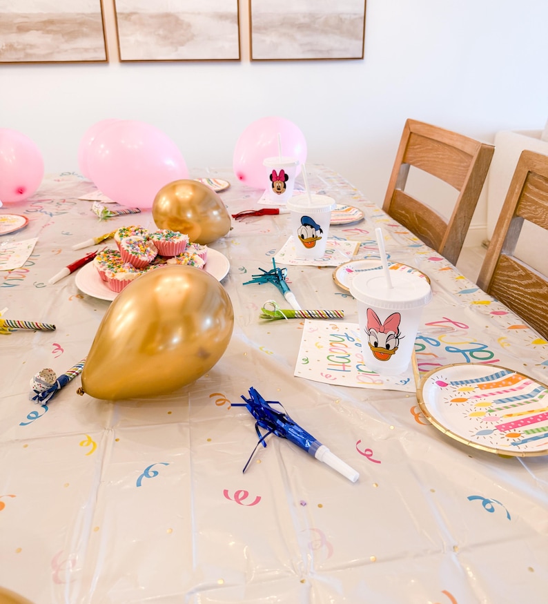 Puede incluir: Una mesa de fiesta con globos rosas y dorados, cupcakes y vasos y platos con personajes. La mesa est&aacute; cubierta con un mantel de pl&aacute;stico transparente con confeti colorido y la palabra "Happy".