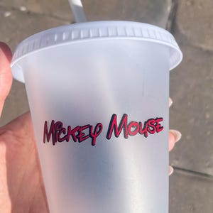 Puede incluir: Un vaso de pl&aacute;stico transparente con tapa y pajita blancas. El vaso presenta las palabras "Mickey Mouse" en letras rojas y negras. La taza est&aacute; dise&ntilde;ada para bebidas fr&iacute;as.
