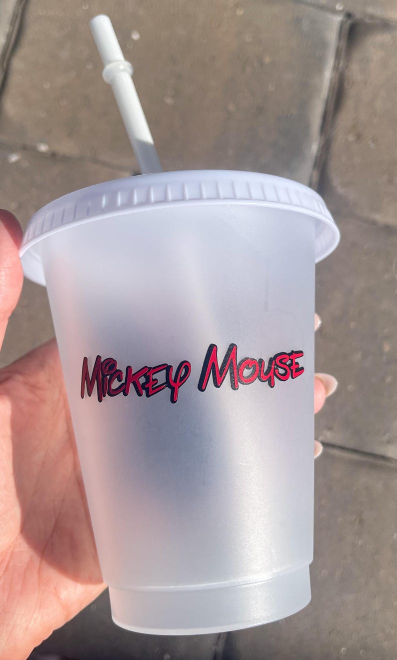 Puede incluir: Un vaso de pl&aacute;stico transparente con tapa y pajita blancas. El vaso presenta las palabras "Mickey Mouse" en letras rojas y negras. La taza est&aacute; dise&ntilde;ada para bebidas fr&iacute;as.