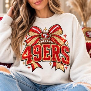 Könnte beinhalten: Ein weißes Sweatshirt mit Rundhalsausschnitt und einer großen Grafik einer rot-goldenen Glitzerschleife, die um einen Football gebunden ist. Der Schriftzug 49ERS ist in großen, roten Glitzerbuchstaben mit Goldrand gedruckt.