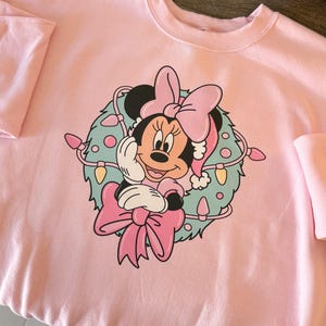 Peut inclure: Sweat-shirt rose clair avec un motif de Minnie Mouse portant un bonnet de P&egrave;re No&euml;l et un n&oelig;ud, entour&eacute;e d'une couronne avec des guirlandes lumineuses. Le sweat-shirt a des manches longues.