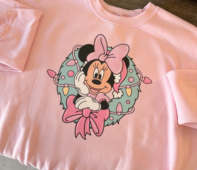 Peut inclure: Sweat-shirt rose clair avec un motif de Minnie Mouse portant un bonnet de P&egrave;re No&euml;l et un n&oelig;ud, entour&eacute;e d'une couronne avec des guirlandes lumineuses. Le sweat-shirt a des manches longues.
