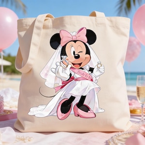 May include: Beige tote bag featuring a bride-themed Minnie Mouse graphic. Minnie Mouse is wearing a white wedding dress, veil, and pink bow. She is holding a ring in each hand. The sash reads "Bride-to-be". Pink balloons and flowers are in the background.