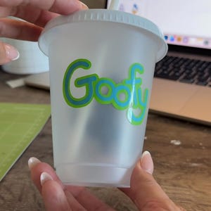 Puede incluir: Un vaso de pl&aacute;stico transparente con tapa blanca y pajita. La palabra "Goofy" est&aacute; impresa en la parte delantera en verde con un contorno azul. El vaso lo sostiene una persona.