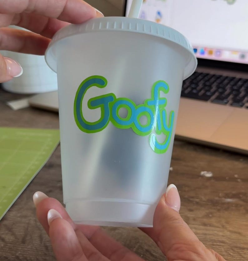 Puede incluir: Un vaso de pl&aacute;stico transparente con tapa blanca y pajita. La palabra "Goofy" est&aacute; impresa en la parte delantera en verde con un contorno azul. El vaso lo sostiene una persona.