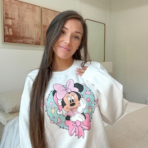 Peut inclure: Sweat-shirt blanc avec un motif de couronne de No&euml;l Minnie Mouse. La couronne comprend un n&oelig;ud rose, un bonnet de P&egrave;re No&euml;l et des accents bleu clair et vert. Le design est centr&eacute; sur le devant.