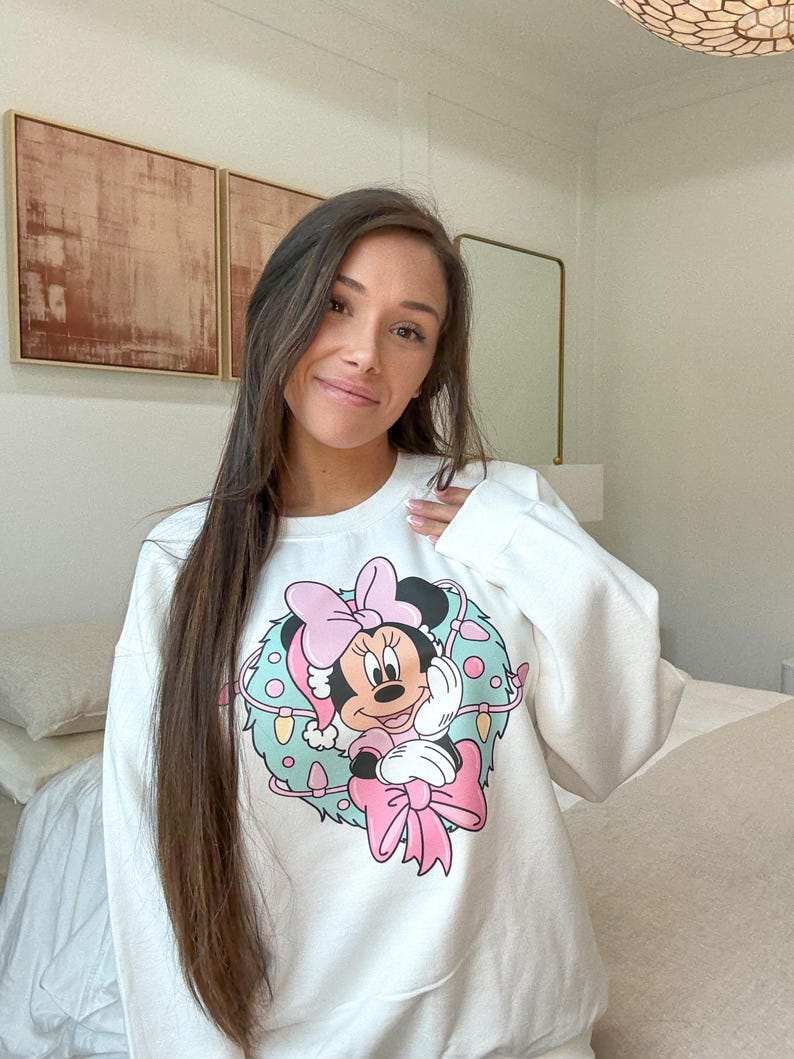 Peut inclure: Sweat-shirt blanc avec un motif de couronne de No&euml;l Minnie Mouse. La couronne comprend un n&oelig;ud rose, un bonnet de P&egrave;re No&euml;l et des accents bleu clair et vert. Le design est centr&eacute; sur le devant.