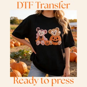 Könnte beinhalten: Schwarzes T-Shirt mit einem Halloween-Motiv, das zwei Teddybären in Kürbiskostümen zeigt. Der orangefarbene Text lautet "DTF Transfer" und "Ready to press". Der Hintergrund zeigt einen Kürbisgarten.
